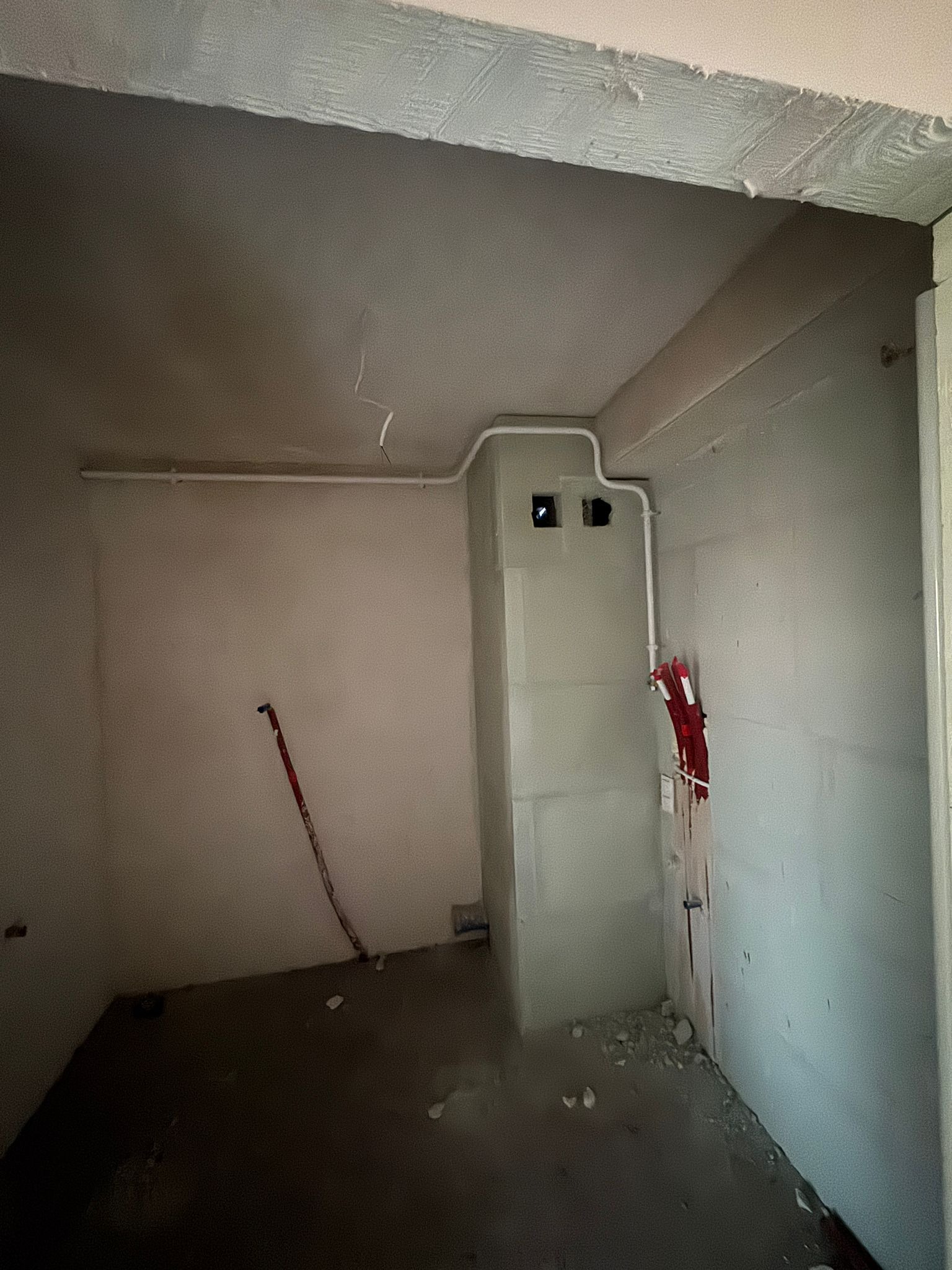 Wnętrze w trakcie prac hydraulicznych: widoczne białe rury instalacji wodnej prowadzone wzdłuż ściany i pod sufitem, z fragmentami czerwonych elementów mocujących, ściany w stanie surowym, podłoga...