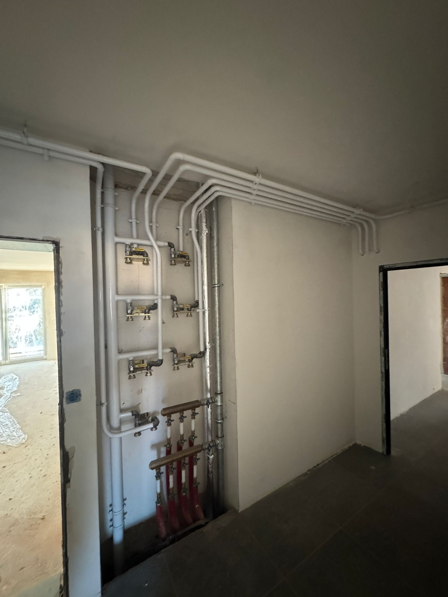 Rozbudowana instalacja grzewcza w surowym wnętrzu, widoczne białe rury, rozdzielacze z zaworami i czerwone pętle grzewcze.