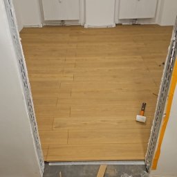 Remonty i wykończenia Maciej Mauer - Układanie paneli podłogowych w trakcie remontu, widoczne narzędzia, deski i fragment ułożonej podłogi w jasnym odcieniu drewna, dwa okna w tle.