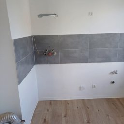 Remonty i wykończenia Maciej Mauer - Wnętrze kuchni w trakcie remontu: widoczne szare płytki na ścianie, przygotowane instalacje elektryczne i wodne, drewnopodobna podłoga, białe ściany i wentylacja.