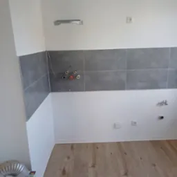 Wnętrze kuchni w trakcie remontu: widoczne szare płytki na ścianie, przygotowane instalacje elektryczne i wodne, drewnopodobna podłoga, białe ściany i wentylacja.