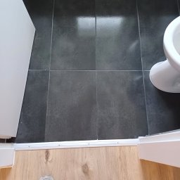 Remonty i wykończenia Maciej Mauer - Ciemne, kwadratowe płytki podłogowe w łazience, widoczne od góry, z fragmentem toalety i progiem łączącym z panelami podłogowymi, na których stoi but.