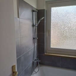Remonty i wykończenia Maciej Mauer - Nowoczesna łazienka po remoncie: szare, podłużne płytki ceramiczne na ścianie, biała wanna, chromowana bateria i deszczownica, okno z szybą ornamentową.