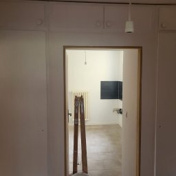 Remonty i wykończenia Maciej Mauer - Wnętrze w trakcie odświeżania: widok przez futrynę na pokój z drabiny malarskiej, grzejnikiem i nową, czarną płytą na ścianie.