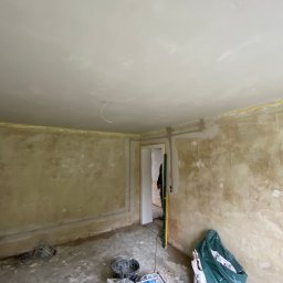 Remonty i wykończenia Maciej Mauer - Pokój w trakcie remontu: przygotowane ściany do malowania, widoczne ślady po szpachlowaniu, worki z materiałami budowlanymi, poziomica oparta o framugę drzwi.
