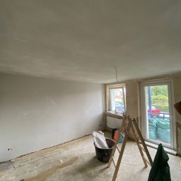 Remonty i wykończenia Maciej Mauer - Pokój w trakcie remontu: ściany po szpachlowaniu, przygotowane do malowania, widoczna drabina, wiadro z zaprawą i worki z materiałami budowlanymi.