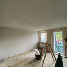 Pokój w trakcie remontu: ściany po szpachlowaniu, przygotowane do malowania, widoczna drabina, wiadro z zaprawą i worki z materiałami budowlanymi.