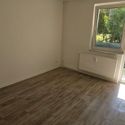 Remonty i wykończenia Maciej Mauer - Pomalowany na biało pokój z drewnopodobną podłogą, białym grzejnikiem i wyjściem na balkon. Świeżo pomalowane ściany w jasnym odcieniu. Na balkonie widoczna osoba w ubraniach roboczych.