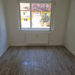 Remonty i wykończenia Maciej Mauer - Pusty pokój po remoncie z panelami podłogowymi, świeżo pomalowanymi ścianami na biało i grzejnikiem pod oknem z widokiem na budynki mieszkalne.