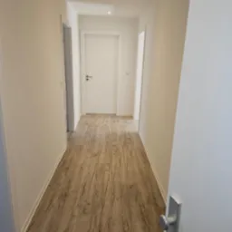 Wnętrze korytarza po remoncie, z drewnopodobną podłogą, białymi ścianami i drzwiami, widoczne listwy przypodłogowe.