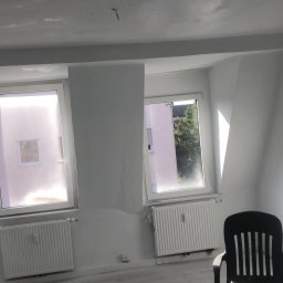 Remonty i wykończenia Maciej Mauer - Pokój w trakcie remontu: surowy strop z widocznymi śladami szpachlowania, dwa okna z grzejnikami, czarne krzesło i maszyna do szlifowania na podłodze.