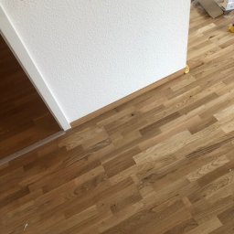 Remonty i wykończenia Maciej Mauer - Fragment pokoju z ułożoną drewnianą podłogą w jodełkę, biała ściana z gniazdkiem elektrycznym i listwą przypodłogową, widoczne opakowania po materiałach budowlanych i fragment białego buta.