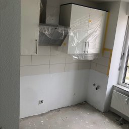 Remonty i wykończenia Maciej Mauer - Wnętrze kuchni w trakcie remontu: białe szafki zabezpieczone folią, świeżo pomalowane ściany, widoczne płytki i przygotowane instalacje, podłoga z plamami farby.