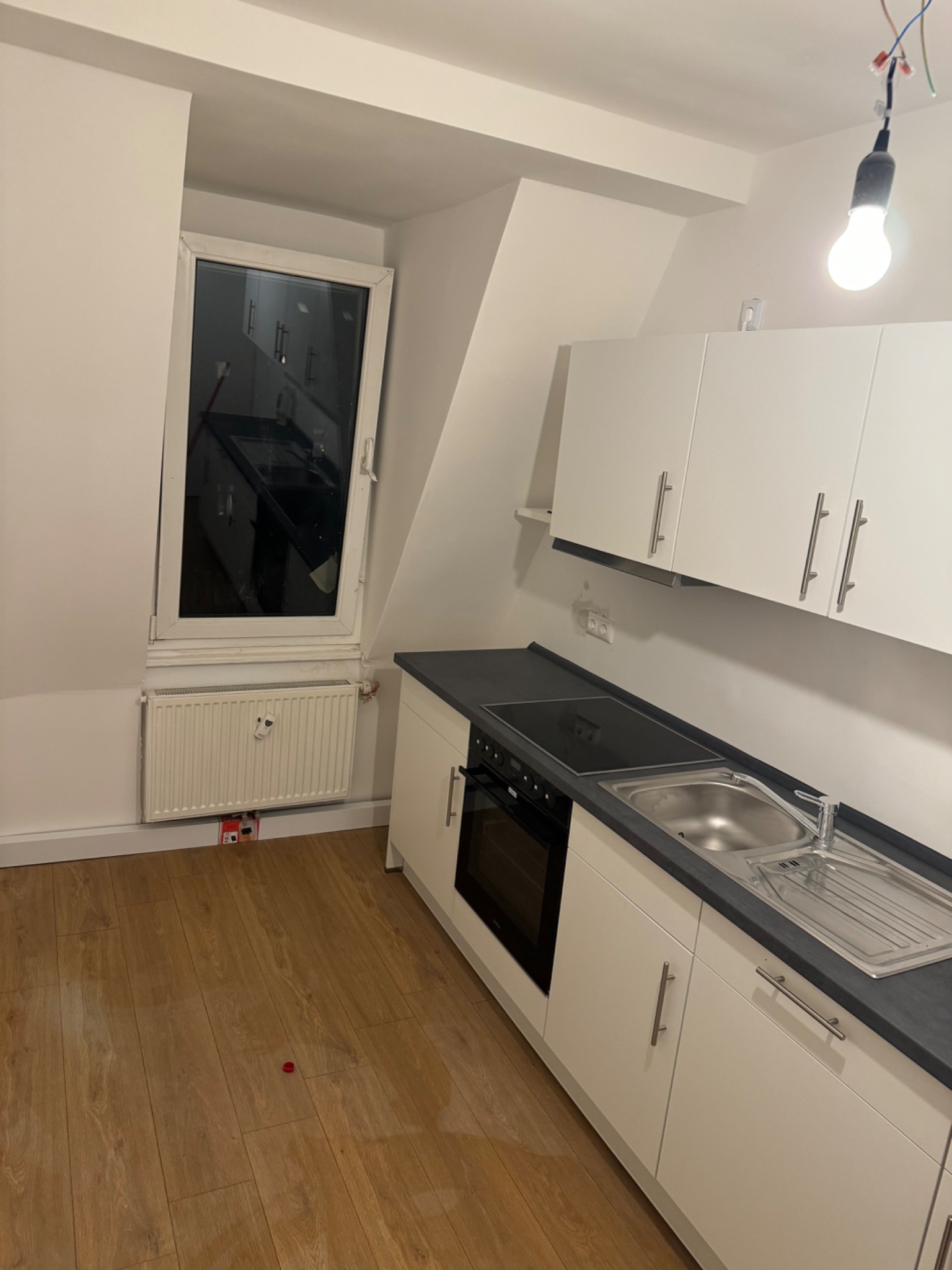 Wnętrze kuchni z białymi szafkami, czarnym blatem i oknem w skosie dachu, widoczny grzejnik pod oknem i wisząca żarówka bez klosza.