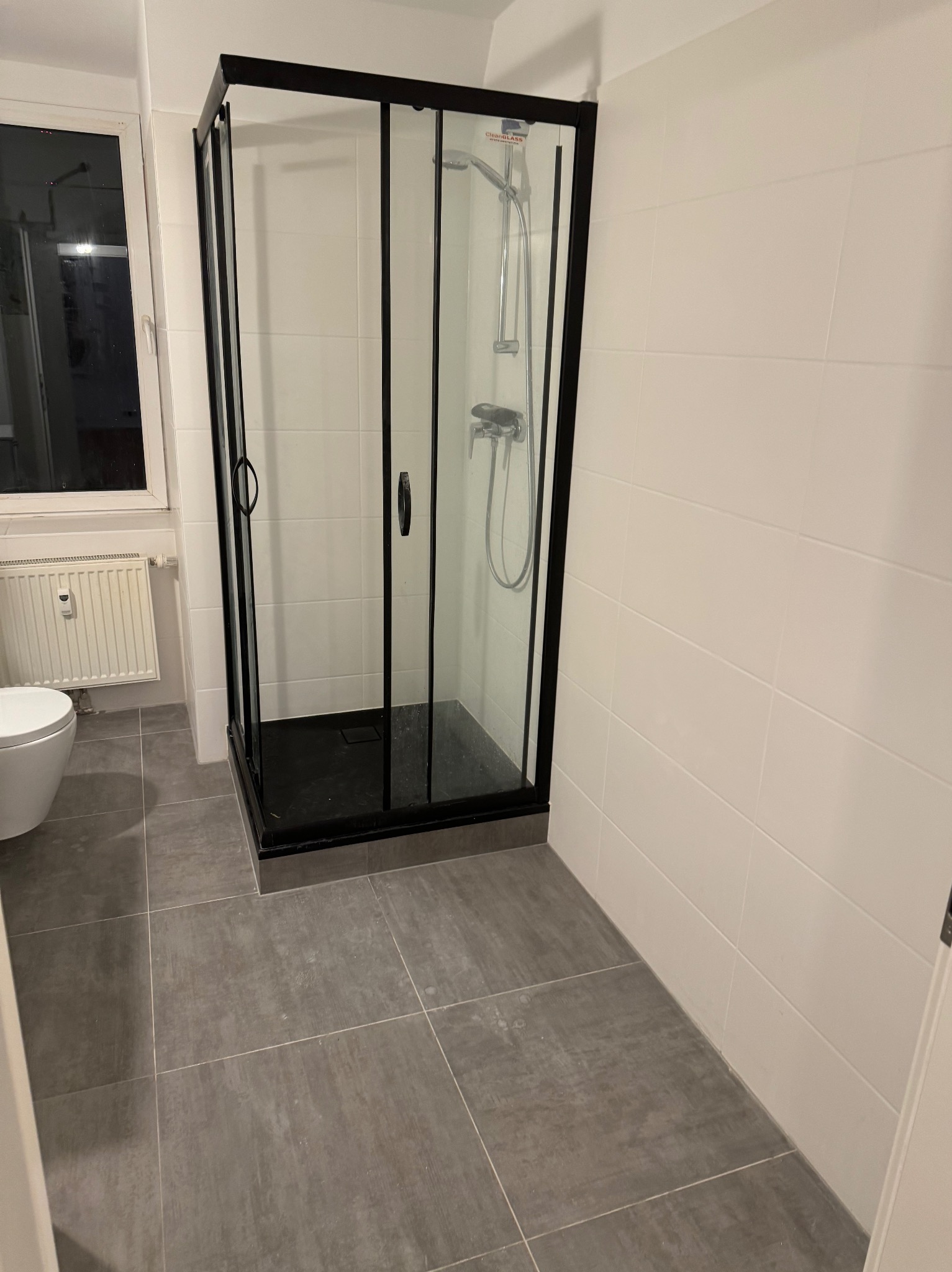 Nowoczesna kabina prysznicowa z czarną ramą i szklanymi drzwiami w łazience z szarymi płytkami podłogowymi i białymi płytkami ściennymi, widoczny fragment toalety i okna.
