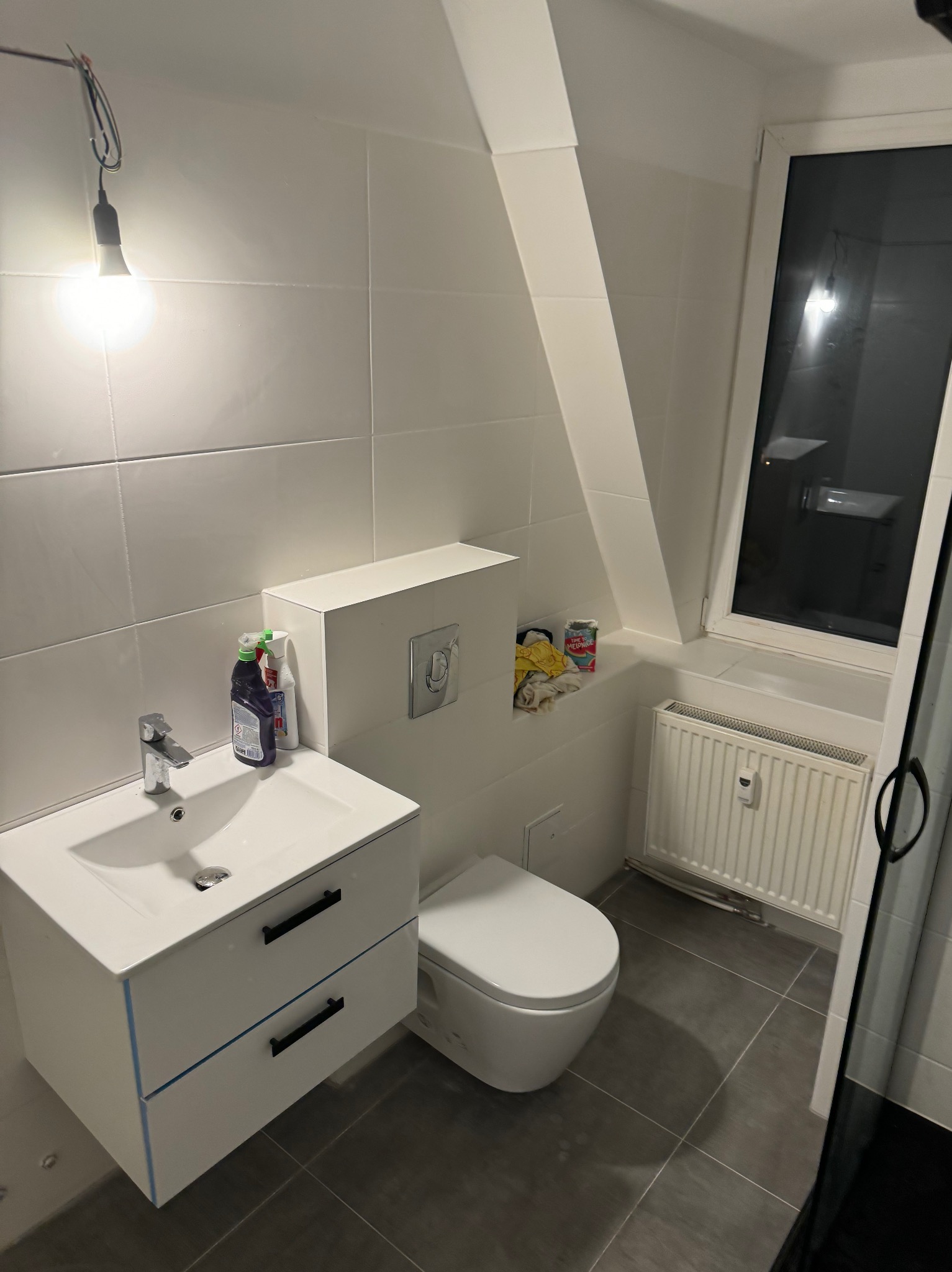 Biała łazienka na poddaszu z umywalką z szafką, toaletą i grzejnikiem, widoczne środki czystości i niedokończone oświetlenie sufitowe.