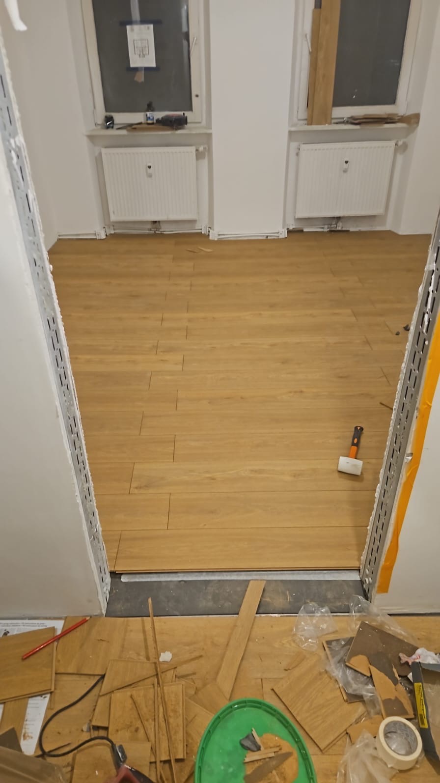 Układanie paneli podłogowych w trakcie remontu, widoczne narzędzia, deski i fragment ułożonej podłogi w jasnym odcieniu drewna, dwa okna w tle.