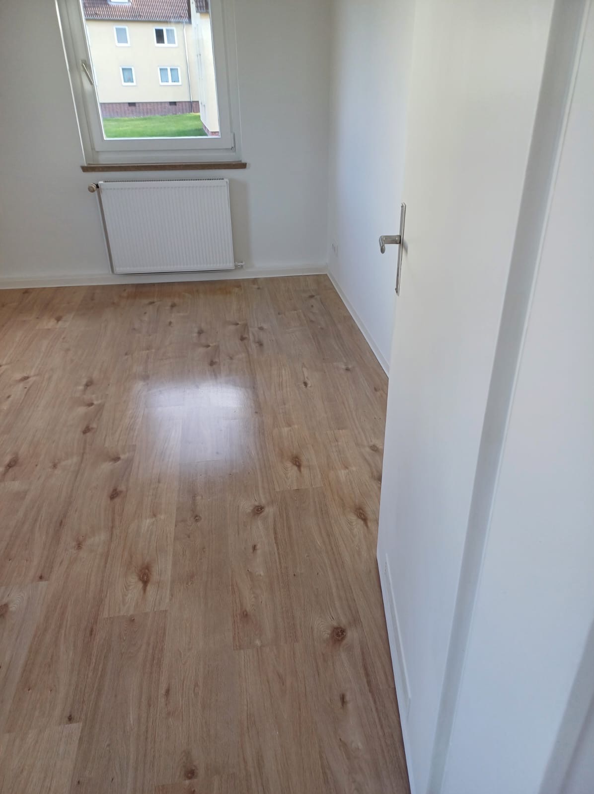 Pusty pokój z panelami podłogowymi w odcieniach brązu, białymi ścianami, grzejnikiem pod oknem i częściowo otwartymi białymi drzwiami z metalową klamką.