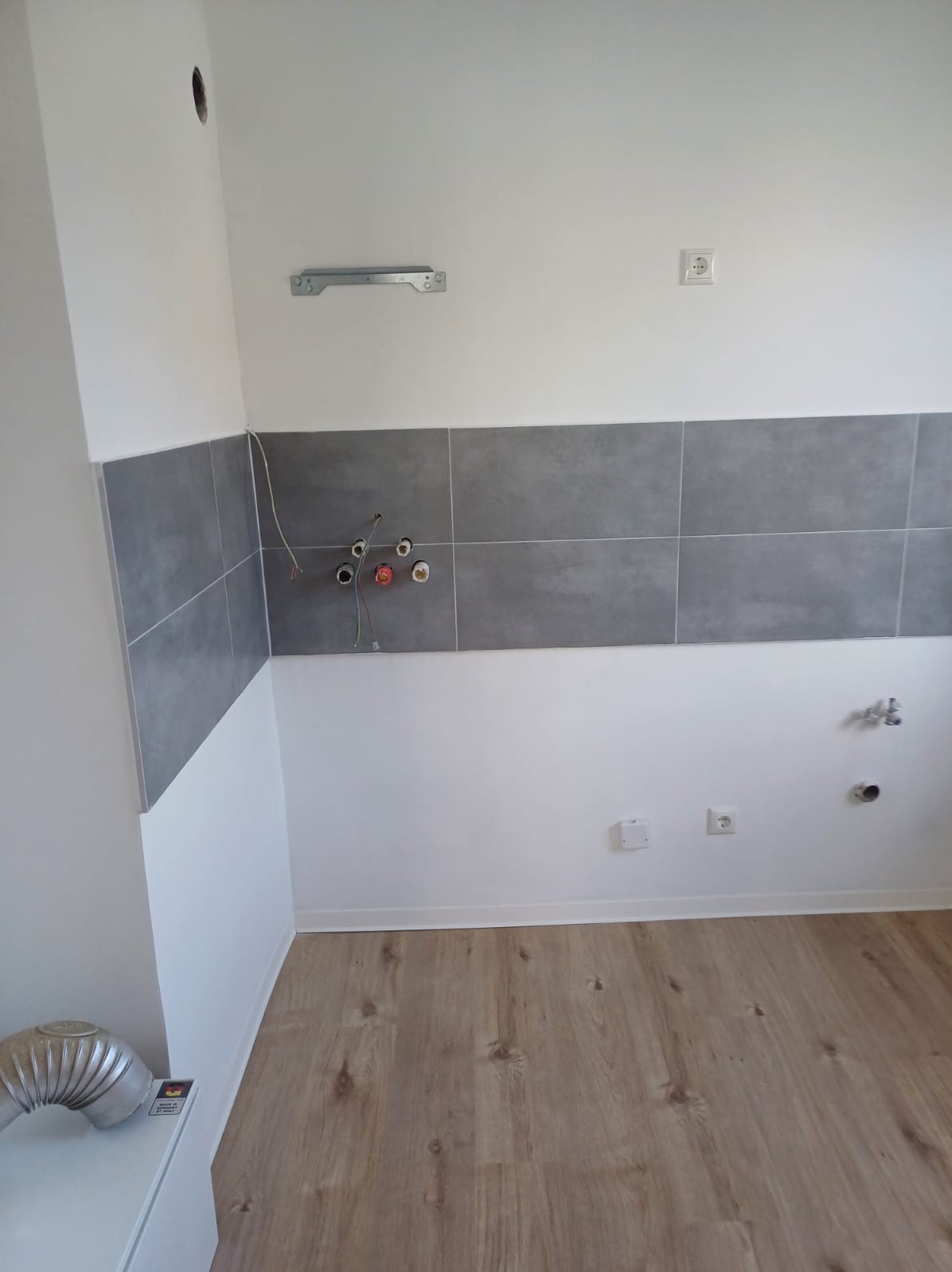 Wnętrze kuchni w trakcie remontu: widoczne szare płytki na ścianie, przygotowane instalacje elektryczne i wodne, drewnopodobna podłoga, białe ściany i wentylacja.