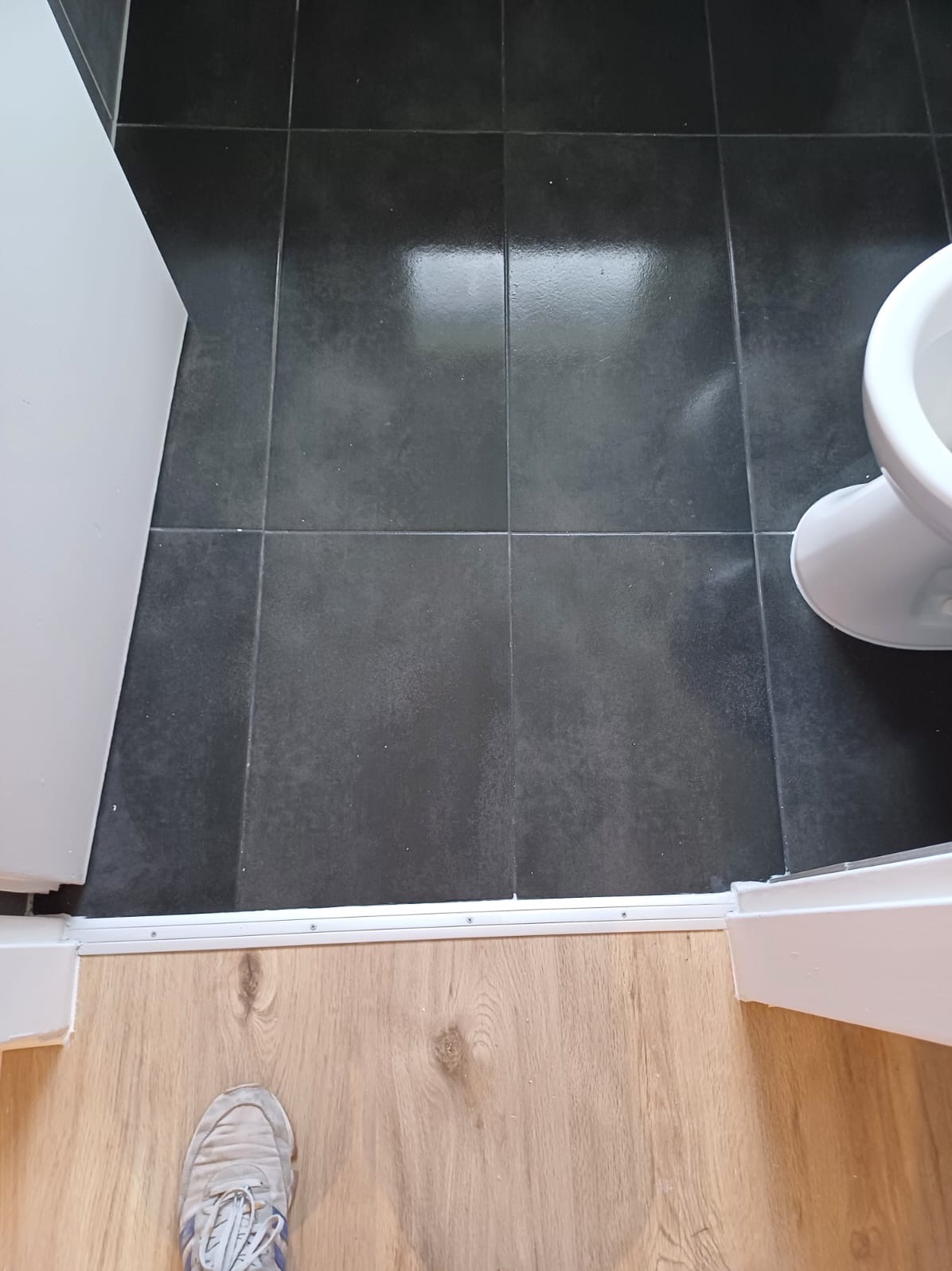 Ciemne, kwadratowe płytki podłogowe w łazience, widoczne od góry, z fragmentem toalety i progiem łączącym z panelami podłogowymi, na których stoi but.