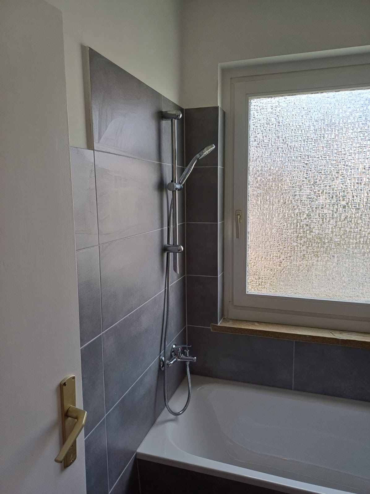 Nowoczesna łazienka po remoncie: szare, podłużne płytki ceramiczne na ścianie, biała wanna, chromowana bateria i deszczownica, okno z szybą ornamentową.