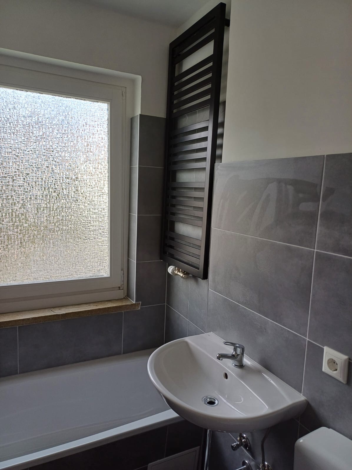 Szara łazienka z wanną, umywalką na postumencie i czarnym grzejnikiem drabinkowym, widoczne szare płytki i okno z szybą ornamentową.