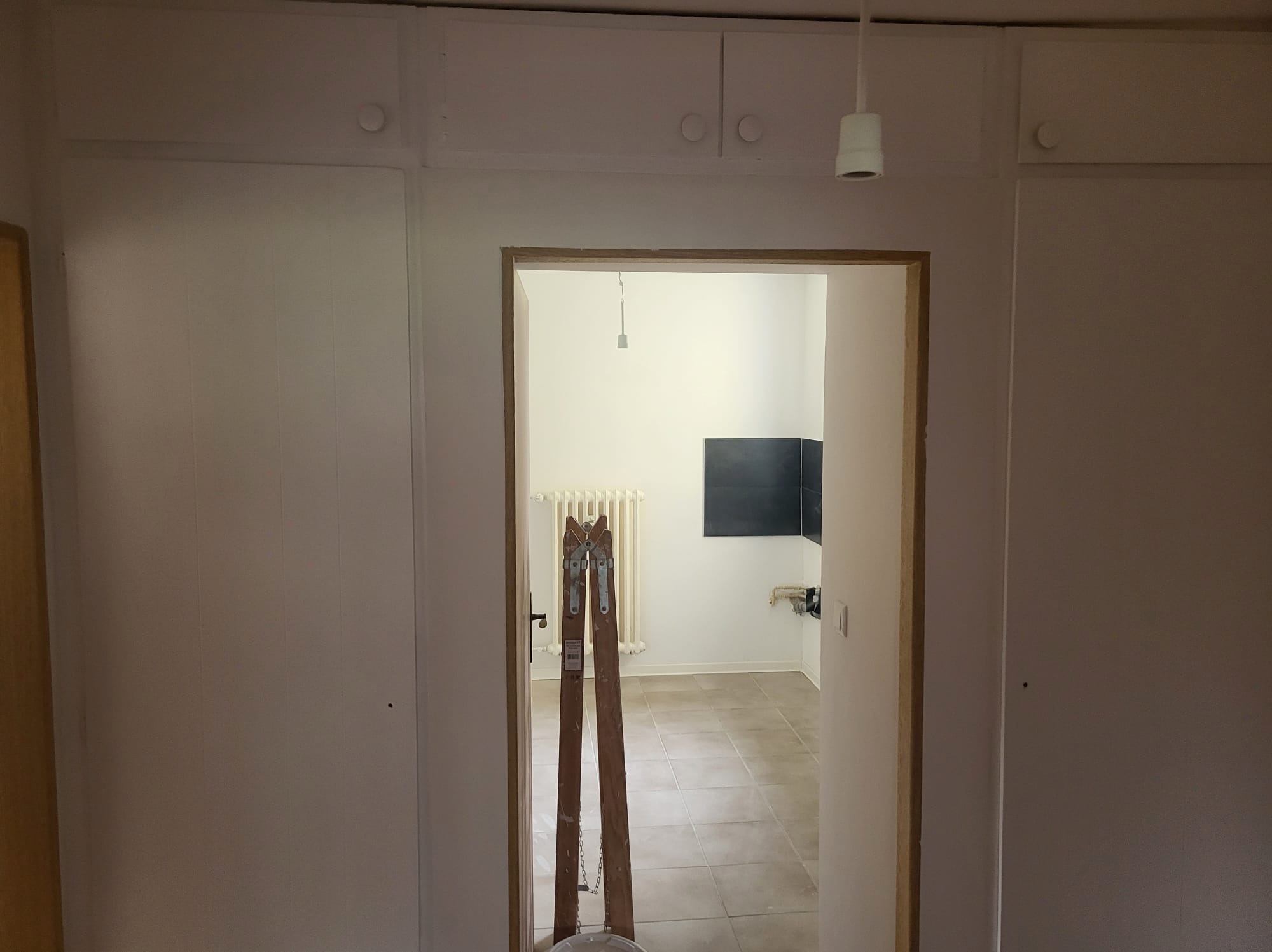 Wnętrze w trakcie odświeżania: widok przez futrynę na pokój z drabiny malarskiej, grzejnikiem i nową, czarną płytą na ścianie.