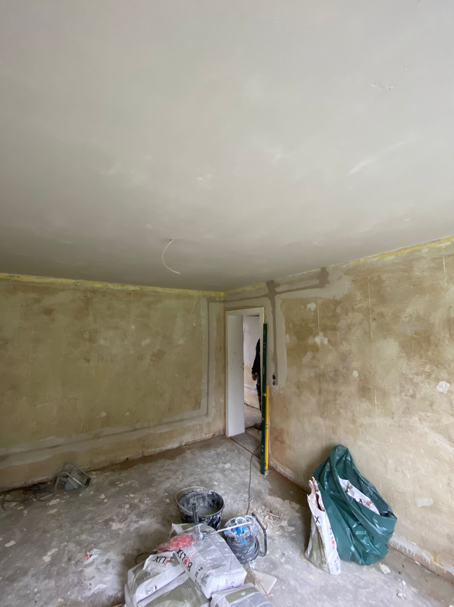 Pokój w trakcie remontu: przygotowane ściany do malowania, widoczne ślady po szpachlowaniu, worki z materiałami budowlanymi, poziomica oparta o framugę drzwi.