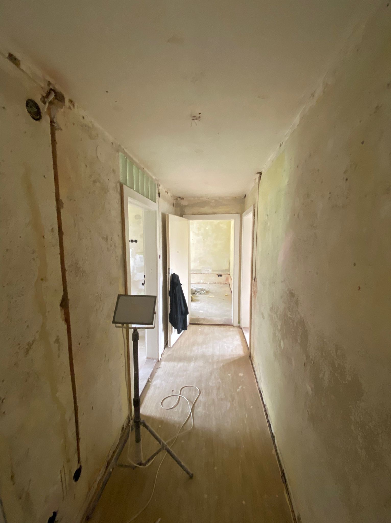 Korytarz w trakcie remontu: ściany z widocznymi śladami po starych powłokach, odsłonięte przewody elektryczne, lampa budowlana na statywie oświetlająca wnętrze, widok na kolejne pomieszczenia w głębi.
