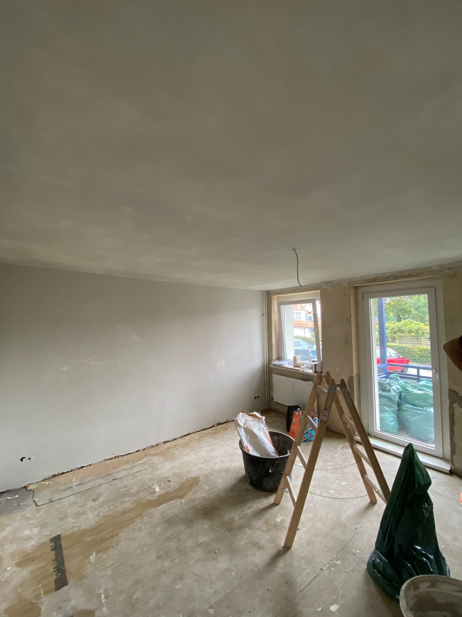 Pokój w trakcie remontu: ściany po szpachlowaniu, przygotowane do malowania, widoczna drabina, wiadro z zaprawą i worki z materiałami budowlanymi.