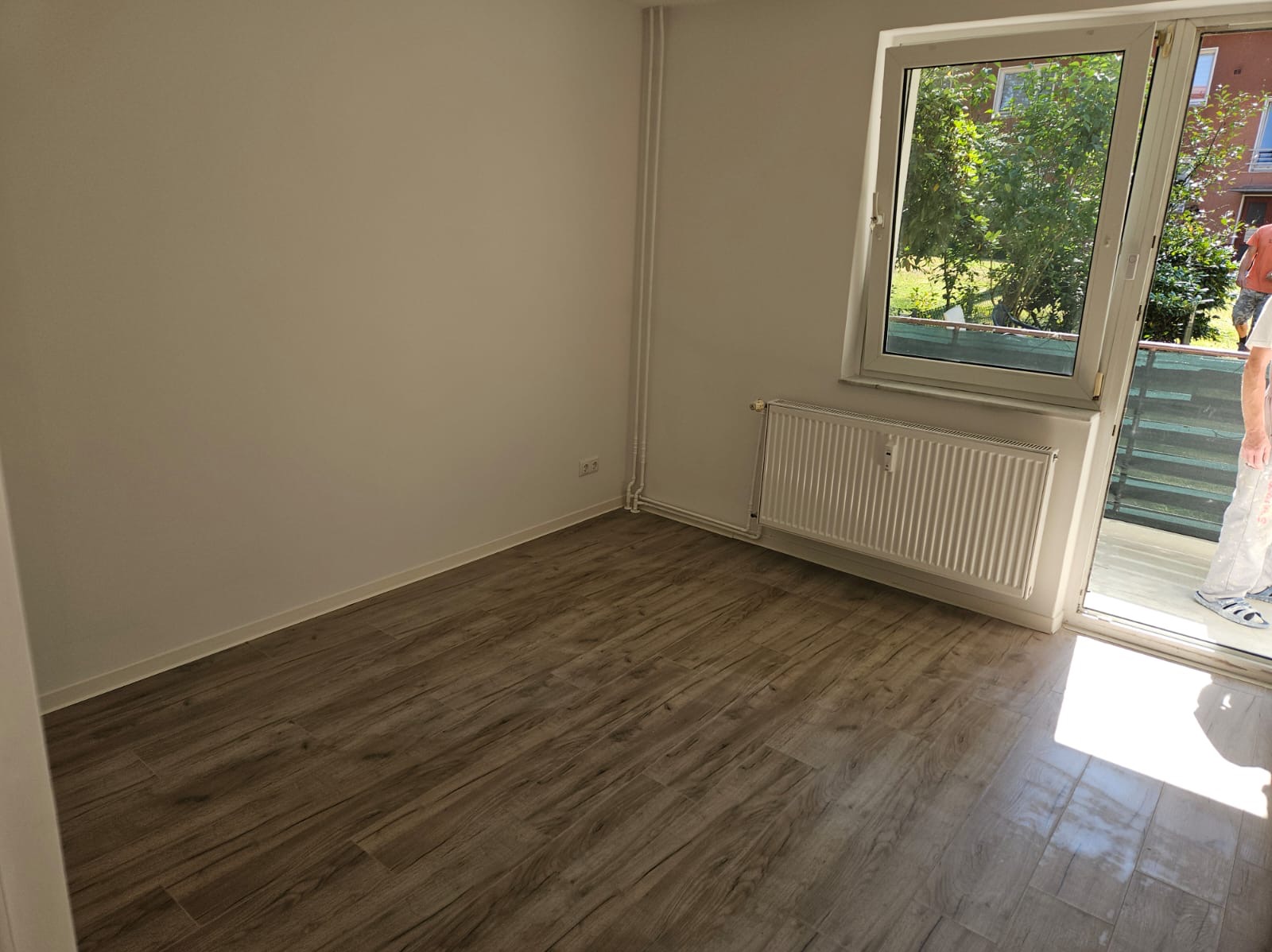 Pomalowany na biało pokój z drewnopodobną podłogą, białym grzejnikiem i wyjściem na balkon. Świeżo pomalowane ściany w jasnym odcieniu. Na balkonie widoczna osoba w ubraniach roboczych.