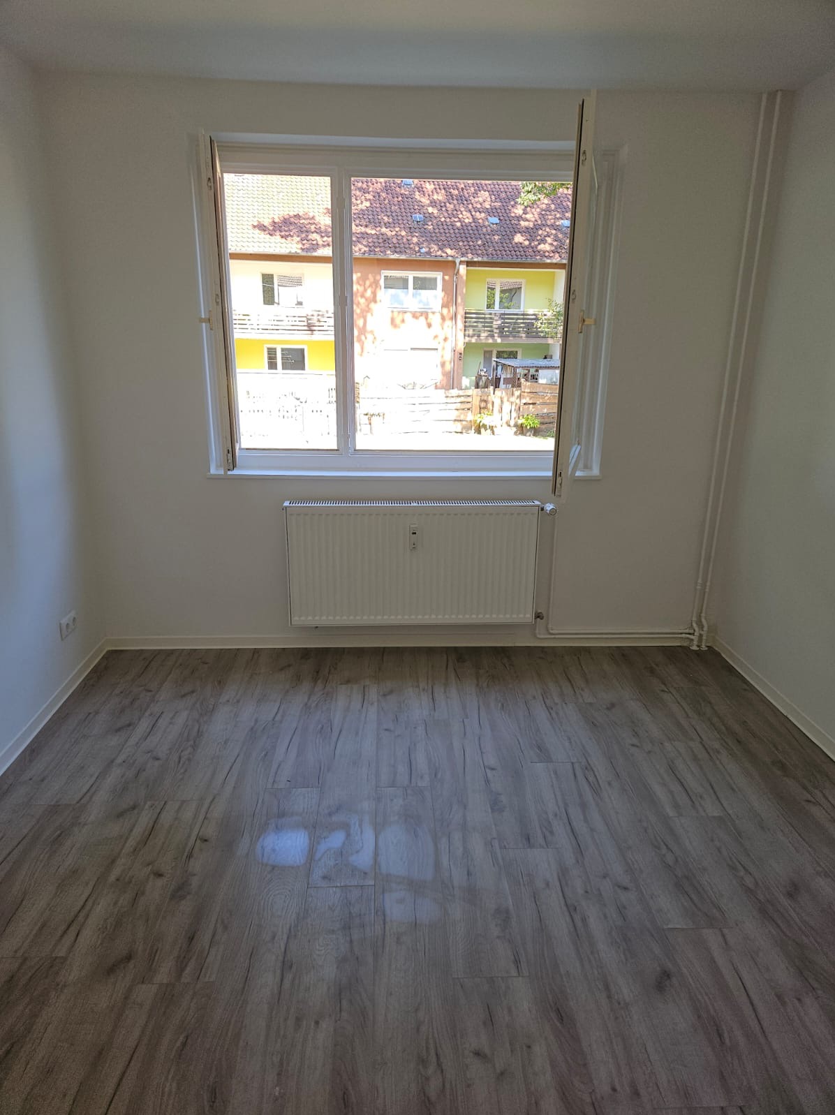 Pusty pokój po remoncie z panelami podłogowymi, świeżo pomalowanymi ścianami na biało i grzejnikiem pod oknem z widokiem na budynki mieszkalne.