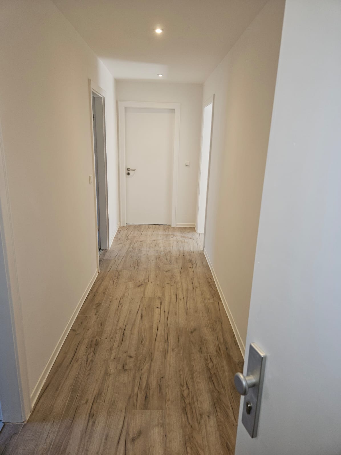 Wnętrze korytarza po remoncie, z drewnopodobną podłogą, białymi ścianami i drzwiami, widoczne listwy przypodłogowe.