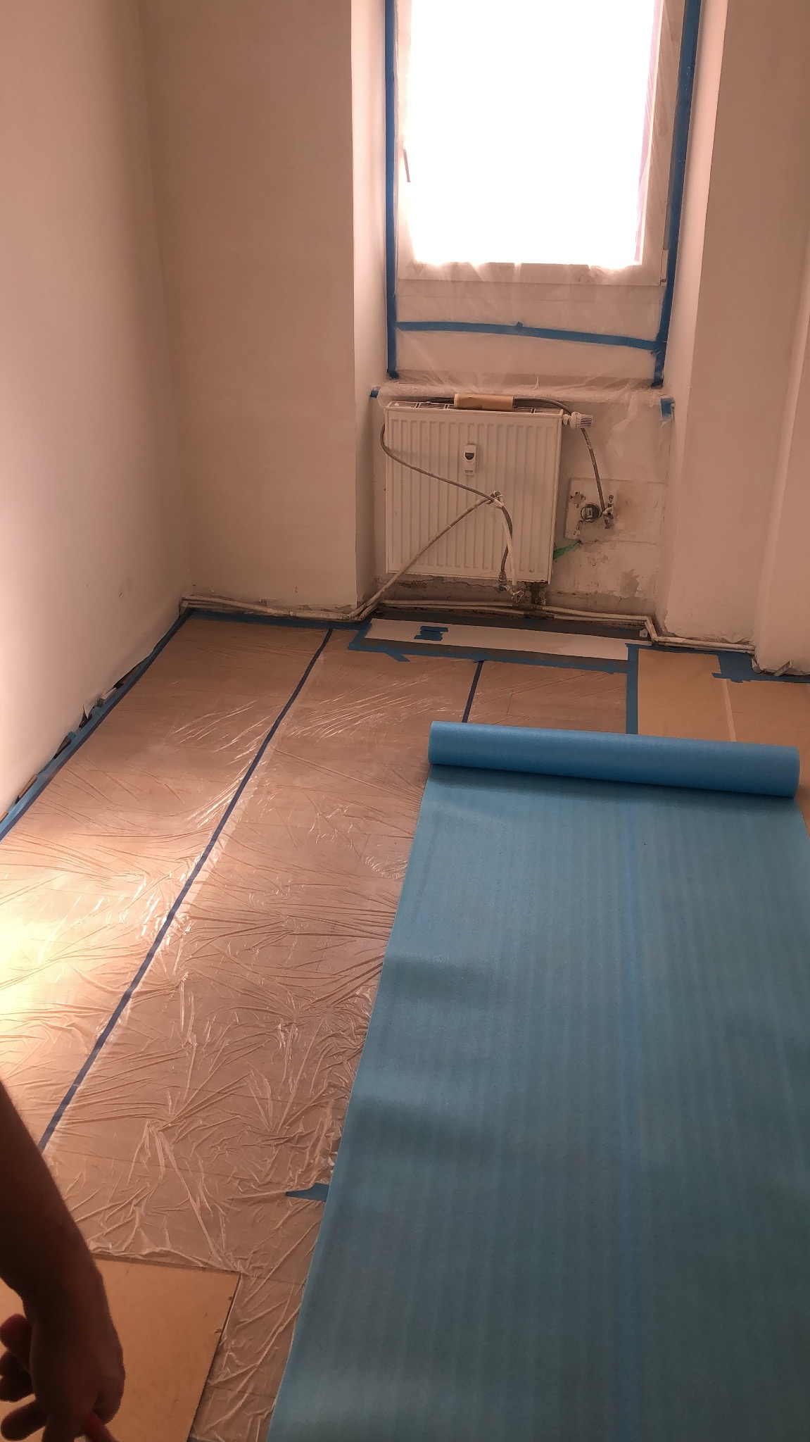 Przygotowanie pokoju do prac wykończeniowych: podłoga zabezpieczona folią malarską i taśmą, okno i grzejnik również zabezpieczone, zwinięta mata ochronna na podłodze.