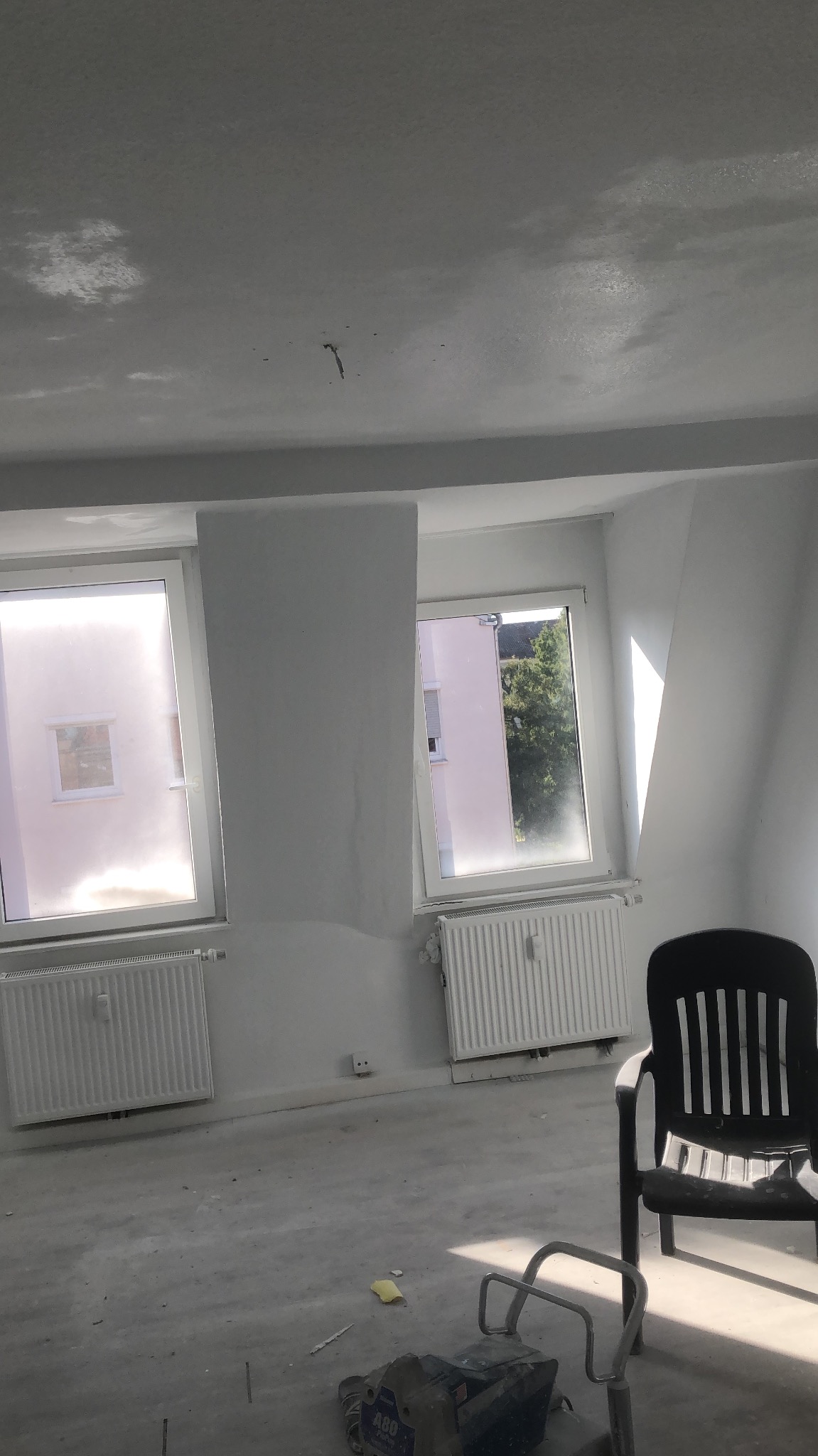 Pokój w trakcie remontu: surowy strop z widocznymi śladami szpachlowania, dwa okna z grzejnikami, czarne krzesło i maszyna do szlifowania na podłodze.