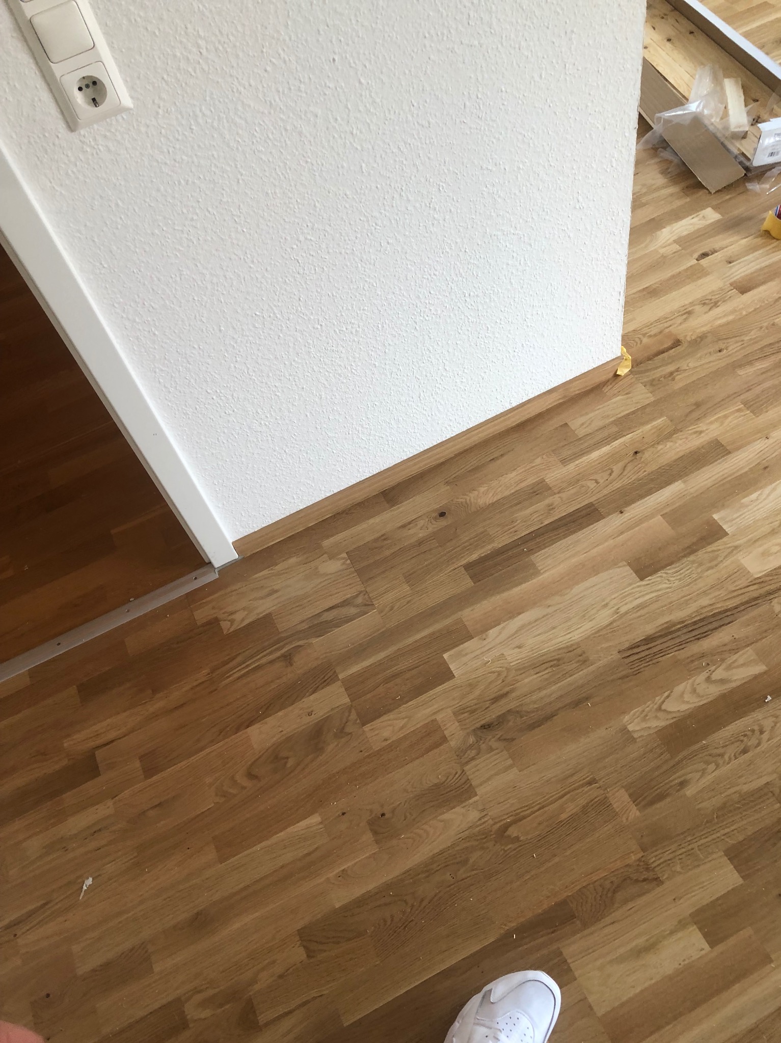 Fragment pokoju z ułożoną drewnianą podłogą w jodełkę, biała ściana z gniazdkiem elektrycznym i listwą przypodłogową, widoczne opakowania po materiałach budowlanych i fragment białego buta.