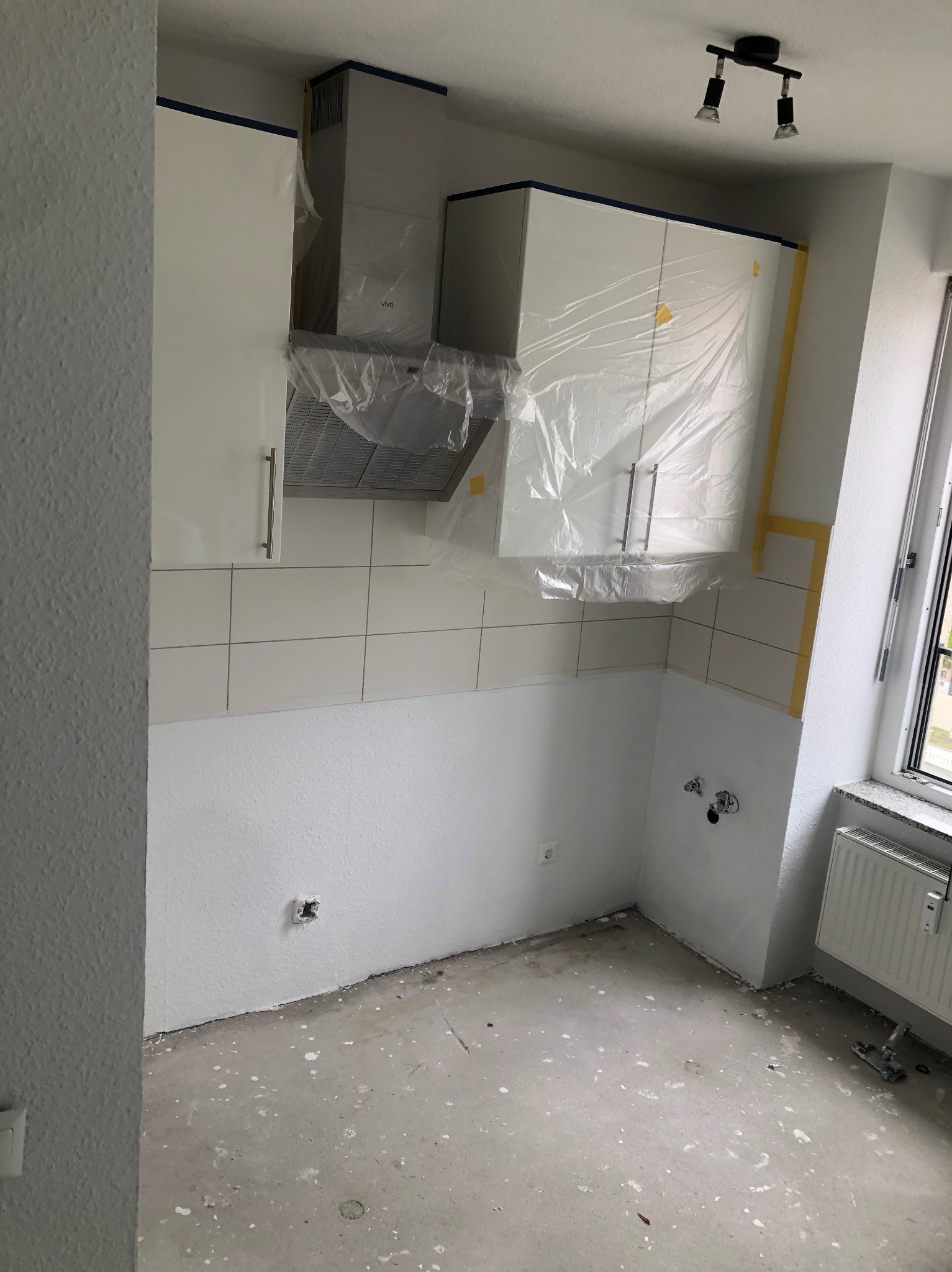 Wnętrze kuchni w trakcie remontu: białe szafki zabezpieczone folią, świeżo pomalowane ściany, widoczne płytki i przygotowane instalacje, podłoga z plamami farby.