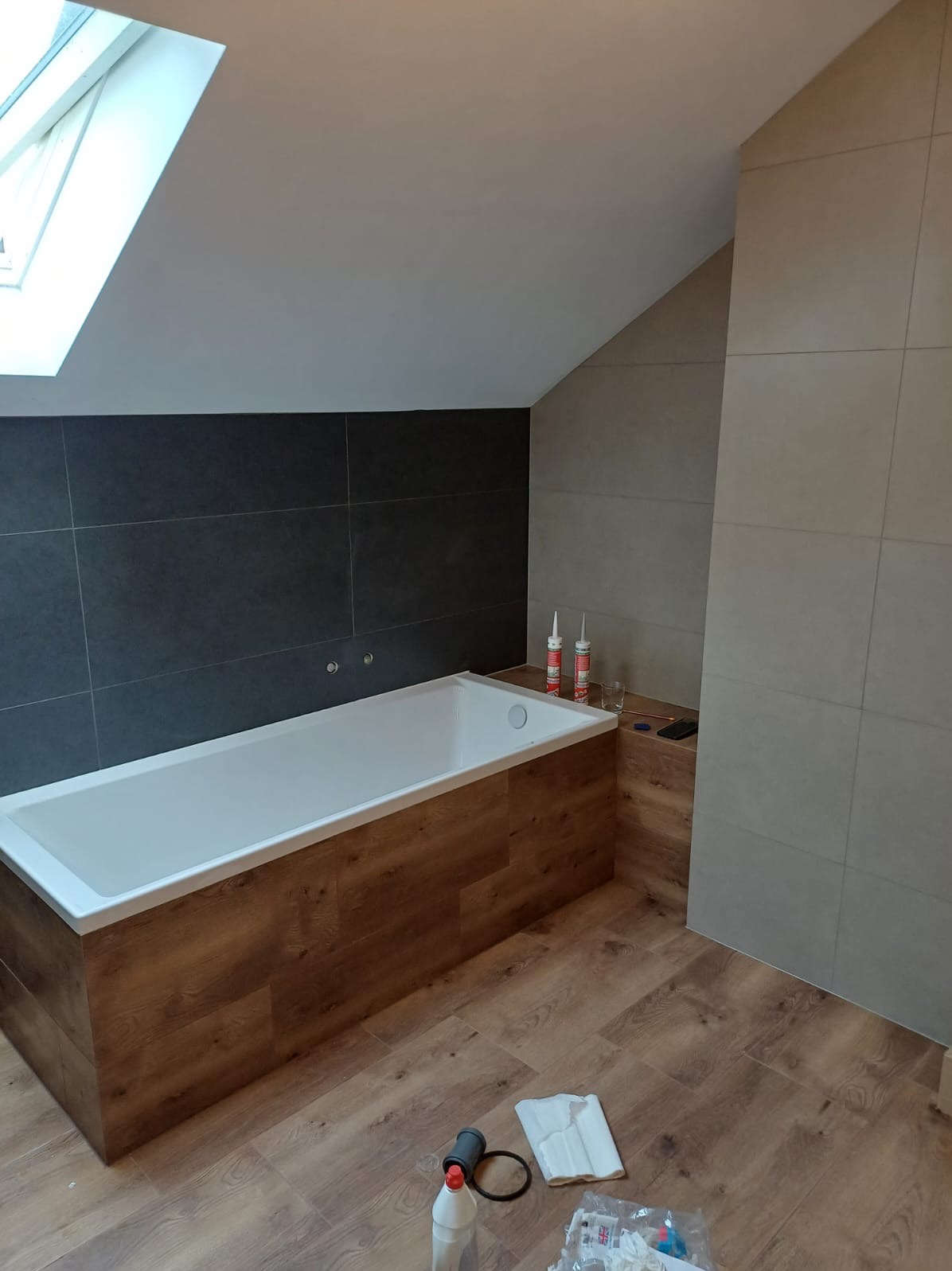 Wnętrze łazienki na poddaszu z białą wanną obudowaną panelami imitującymi drewno, szare płytki na ścianie i akcesoria montażowe na podłodze