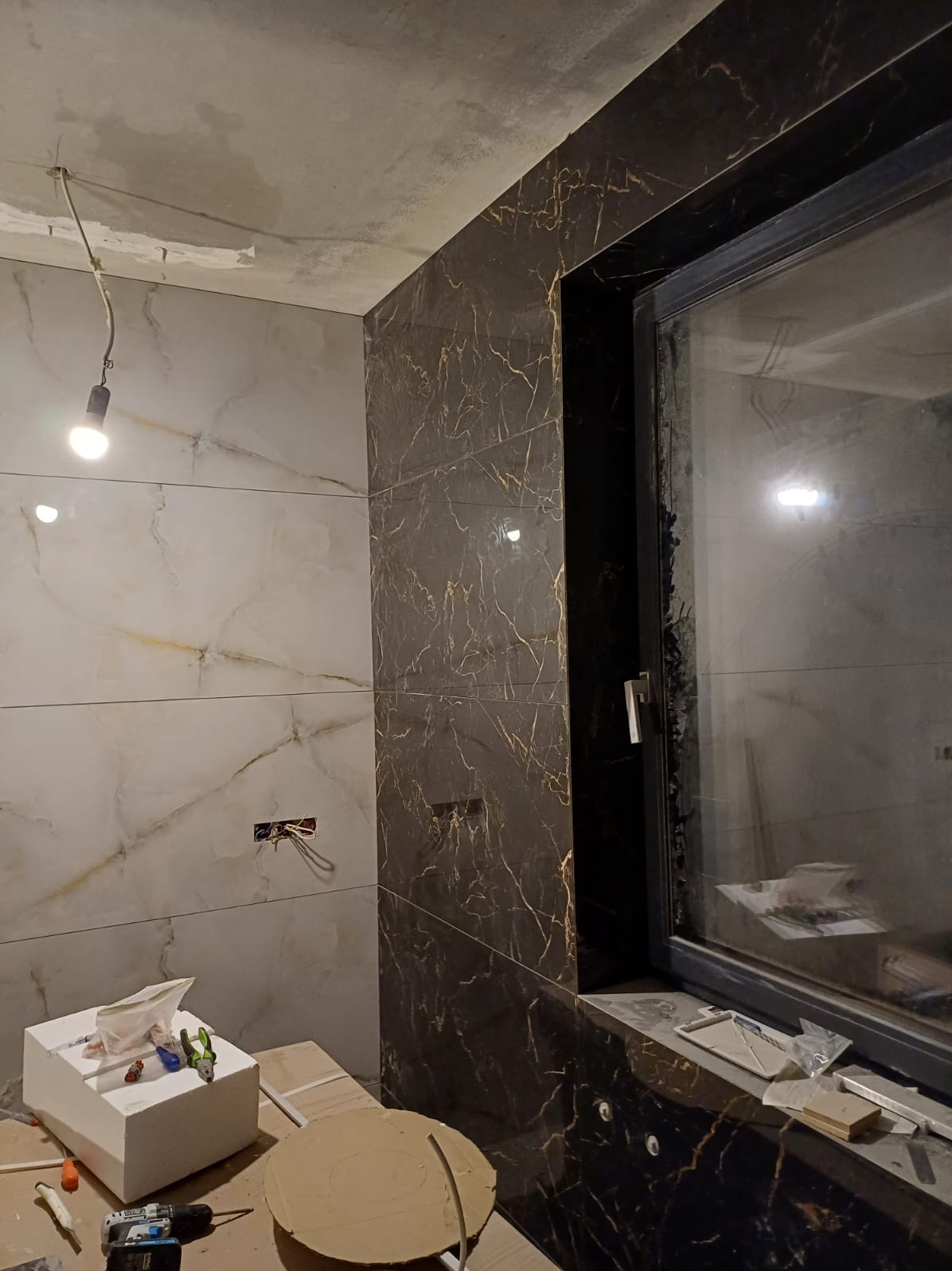Wnętrze łazienki w trakcie remontu, widoczne płytki imitujące marmur w kolorze białym i czarnym ze złotymi żyłami, okno z czarną ramą, narzędzia i materiały budowlane na podłodze, surowy sufit...