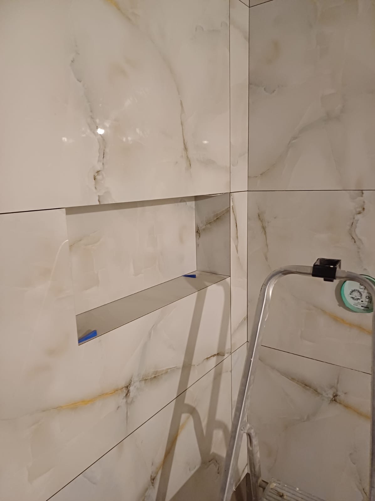 Wykończona ściana łazienki z białych płytek imitujących marmur z wnęką, zabezpieczona niebieską taśmą, widoczny fragment aluminiowej drabiny.