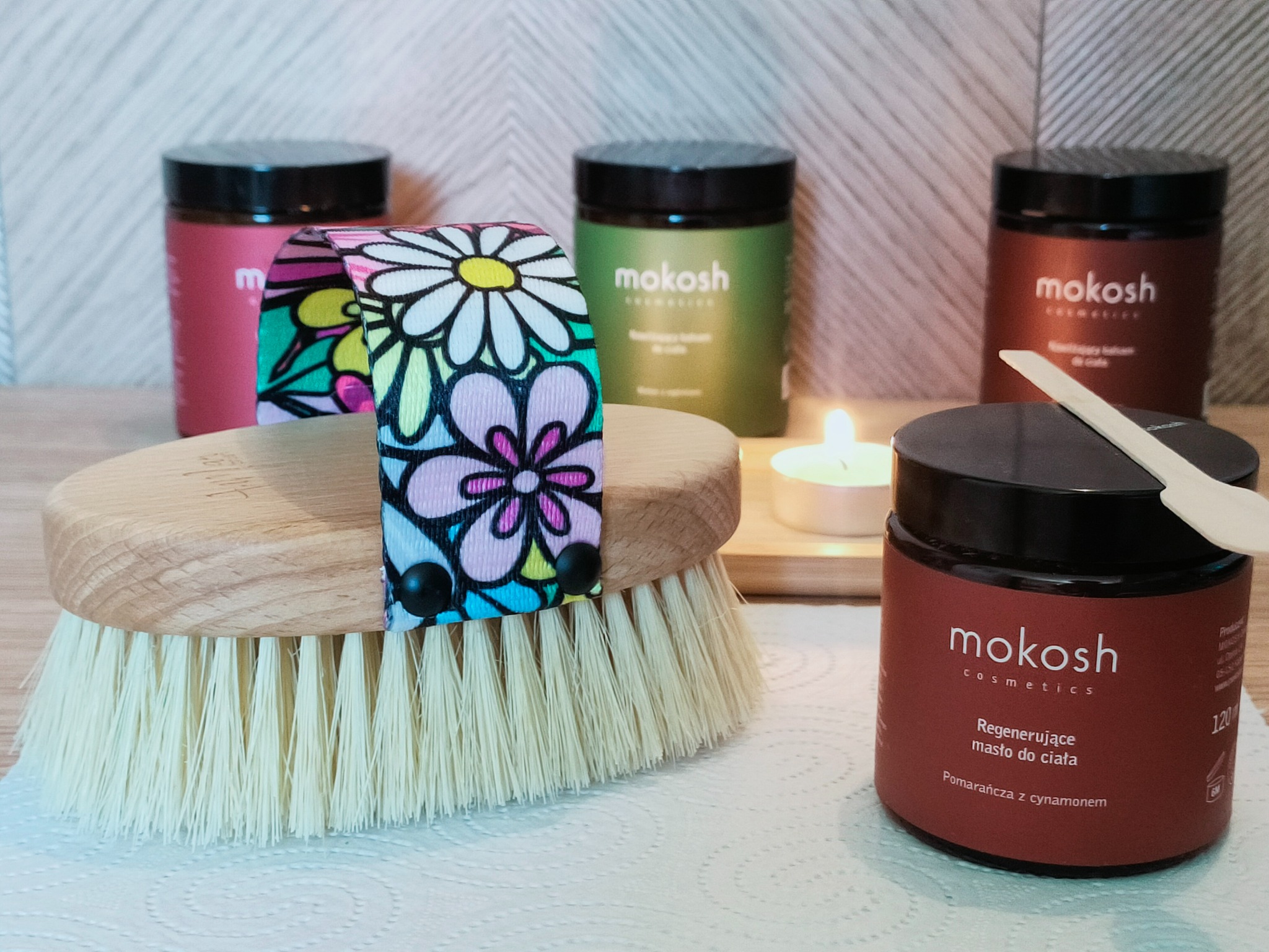 Szczotka do masażu na sucho z kolorową opaską z motywem kwiatowym, obok słoiczki z kosmetykami Mokosh, w tle zapalona świeczka.