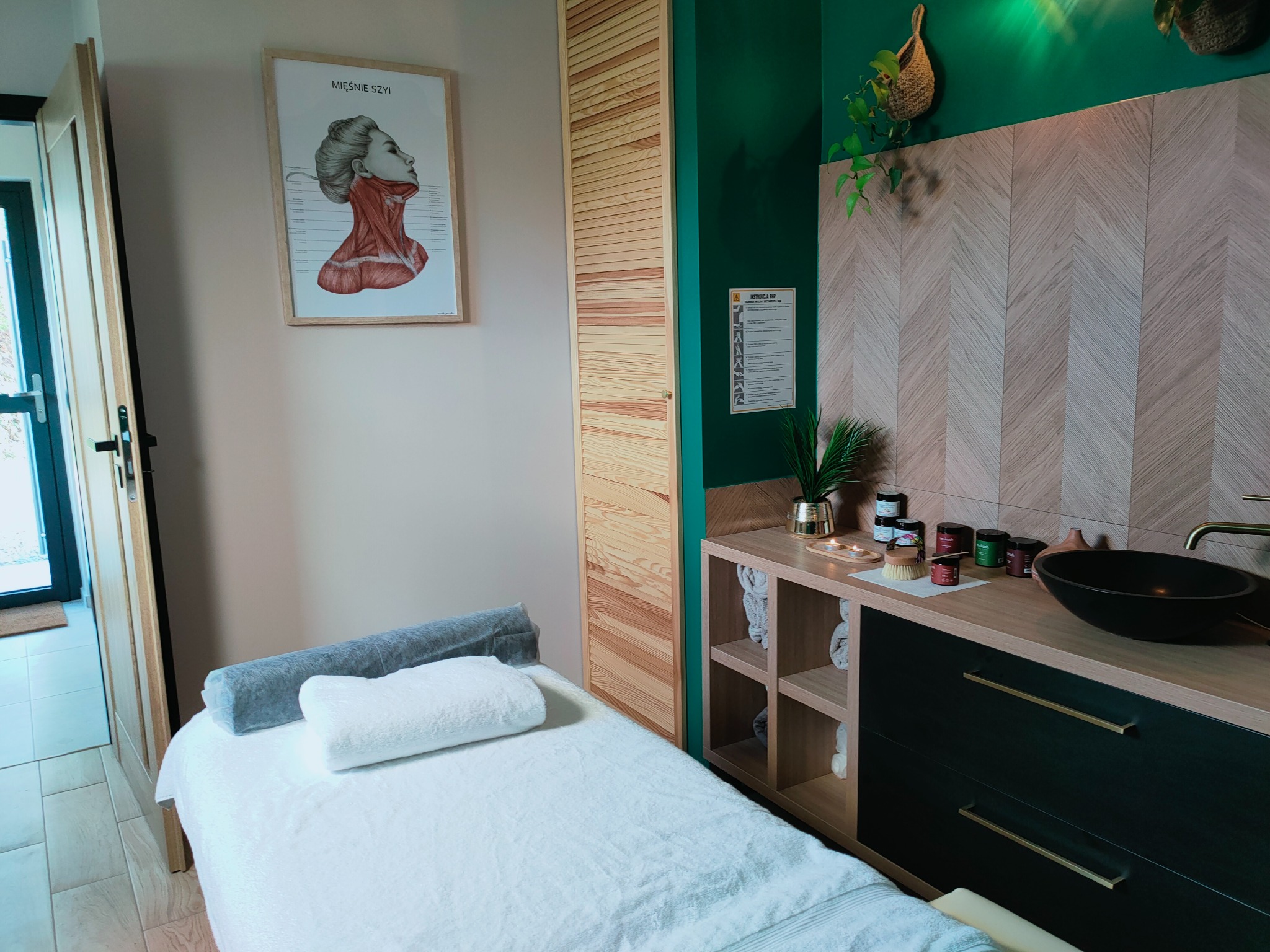 Nowoczesny gabinet masażu z łóżkiem, plakatem anatomicznym mięśni szyi, szafką z kosmetykami i umywalką w kolorach zieleni i drewna.