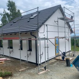 Andrii Matiitsiv - Biały dom jednorodzinny z ciemną dachówką w trakcie prac wykończeniowych elewacji. Widoczne rusztowania, okna z czarnymi ramami i elementy zabezpieczone folią.