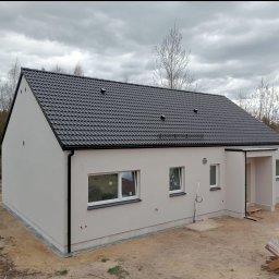 Andrii Matiitsiv - Wykończony dom jednorodzinny z szarą elewacją i ciemną dachówką, widok z boku. Elementy wykończeniowe w kolorze czarnym. Otoczenie w stanie surowym.