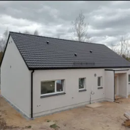 Wykończony dom jednorodzinny z szarą elewacją i ciemną dachówką, widok z boku. Elementy wykończeniowe w kolorze czarnym. Otoczenie w stanie surowym.