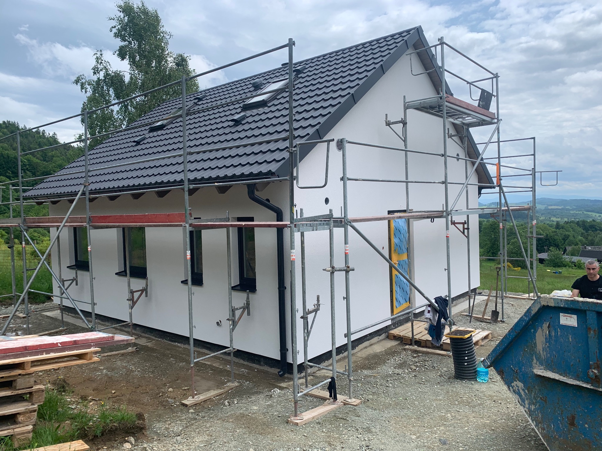 Biały dom jednorodzinny z ciemną dachówką w trakcie prac wykończeniowych elewacji. Widoczne rusztowania, okna z czarnymi ramami i elementy zabezpieczone folią.