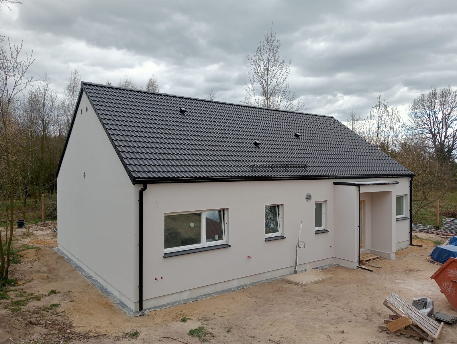 Nowoczesny dom jednorodzinny z jasną elewacją i ciemnym dachem, widoczny z perspektywy ogrodu w pochmurny dzień. Świeżo otynkowana fasada, czarne rynny i okna.