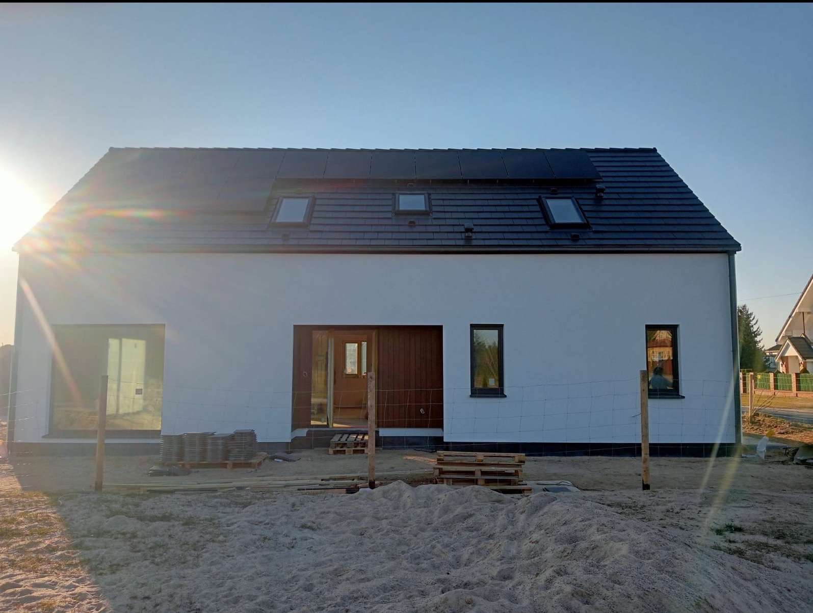 Biała elewacja domu jednorodzinnego z ciemną dachówką, oknami dachowymi i panelami słonecznymi. Częściowo ukończona, widoczne materiały budowlane i ogrodzenie.