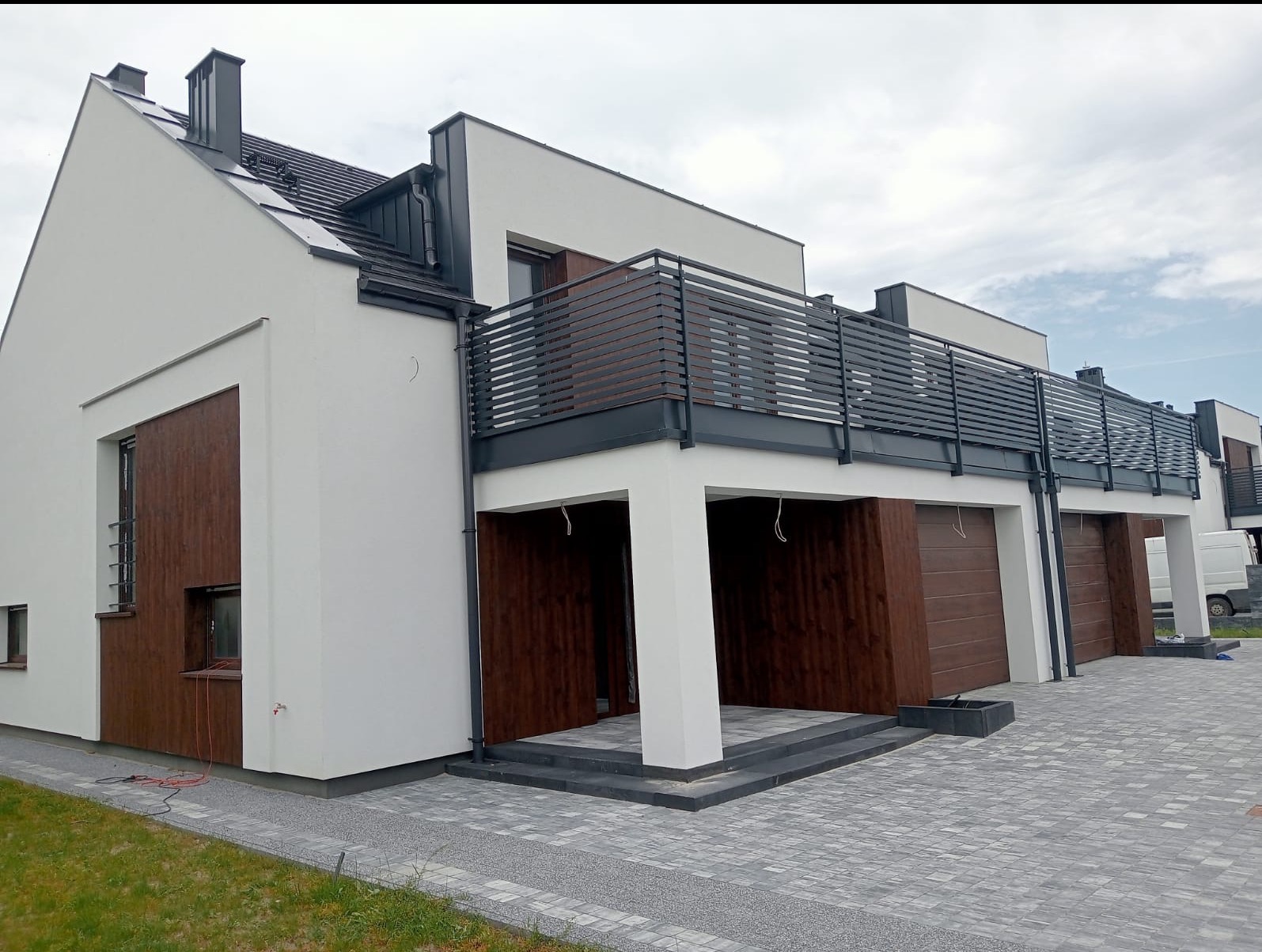 Nowoczesna elewacja szeregowca z drewnianymi panelami i balkonem z metalową balustradą. Jasna kolorystyka ścian i szara kostka brukowa. Widok na kilka segmentów.