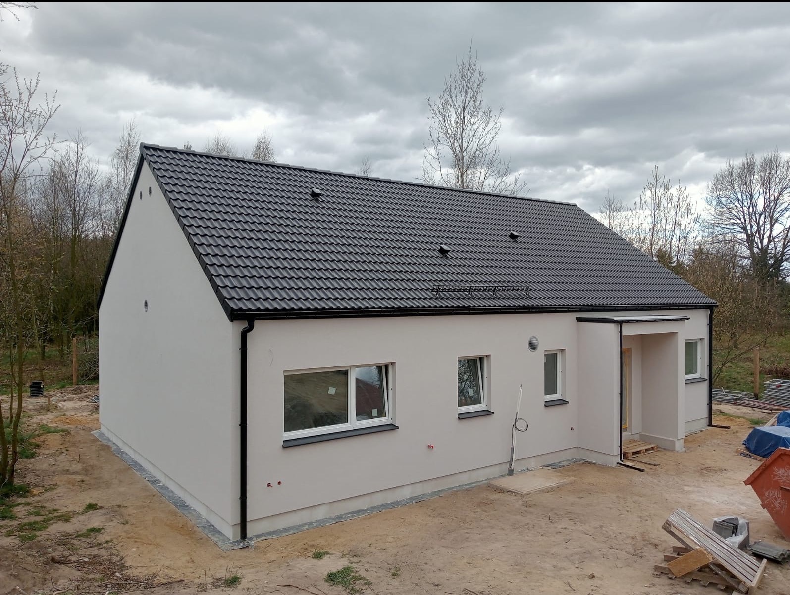 Wykończony dom jednorodzinny z szarą elewacją i ciemną dachówką, widok z boku. Elementy wykończeniowe w kolorze czarnym. Otoczenie w stanie surowym.